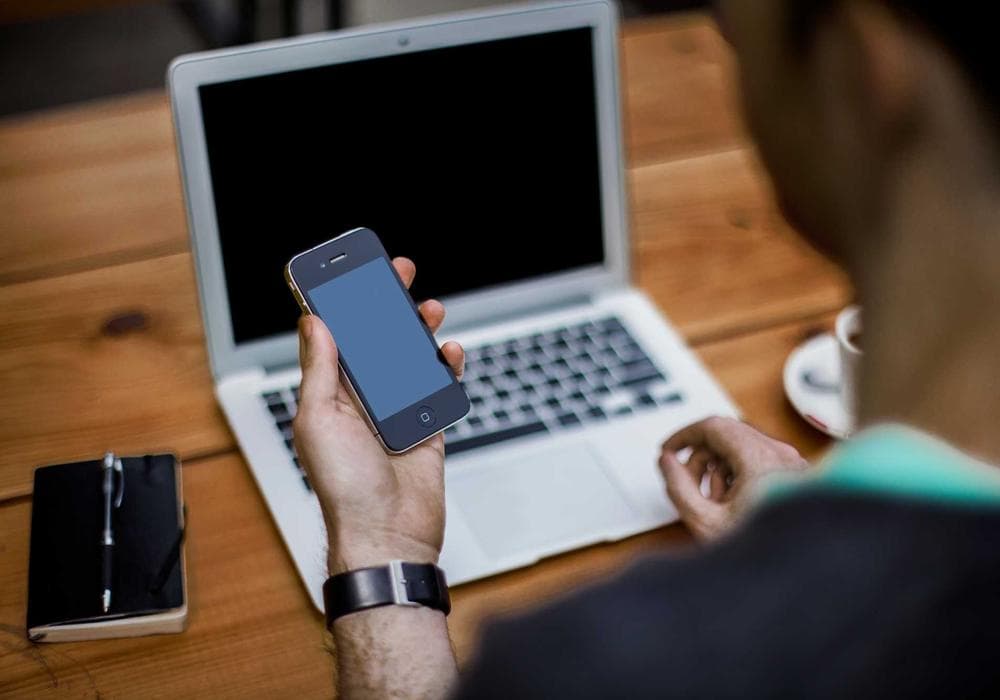 Hands holding a phone above a laptop on a wooden desk